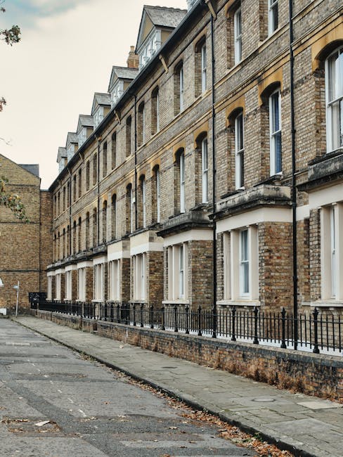 A daytime view of a narrow, curved pathway alongside a canal in an urban residential area. On the left, there is a multi-storey building with yellow brickwork and graffiti-covered walls at ground leve