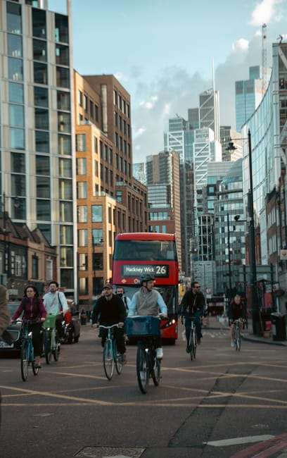 A red double-decker bus displaying the route number 26 and the destination Hackney Wick is positioned in the middle of a busy city street in Hackney Central, surrounded by modern high-rise office buil